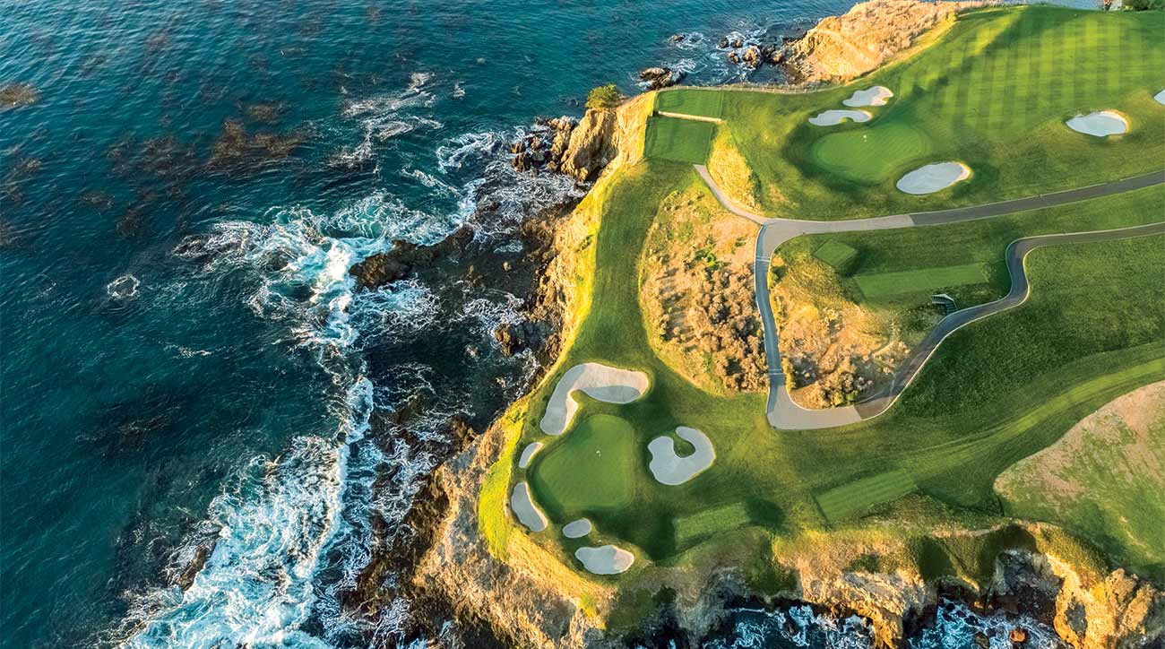 Aerial view of a dramatic coastal golf course with emerald green fairways and white sand bunkers perched atop golden cliffs. Turquoise waters of the Pacific Ocean crash against the rocky coastline below, creating a stunning contrast between land and sea.