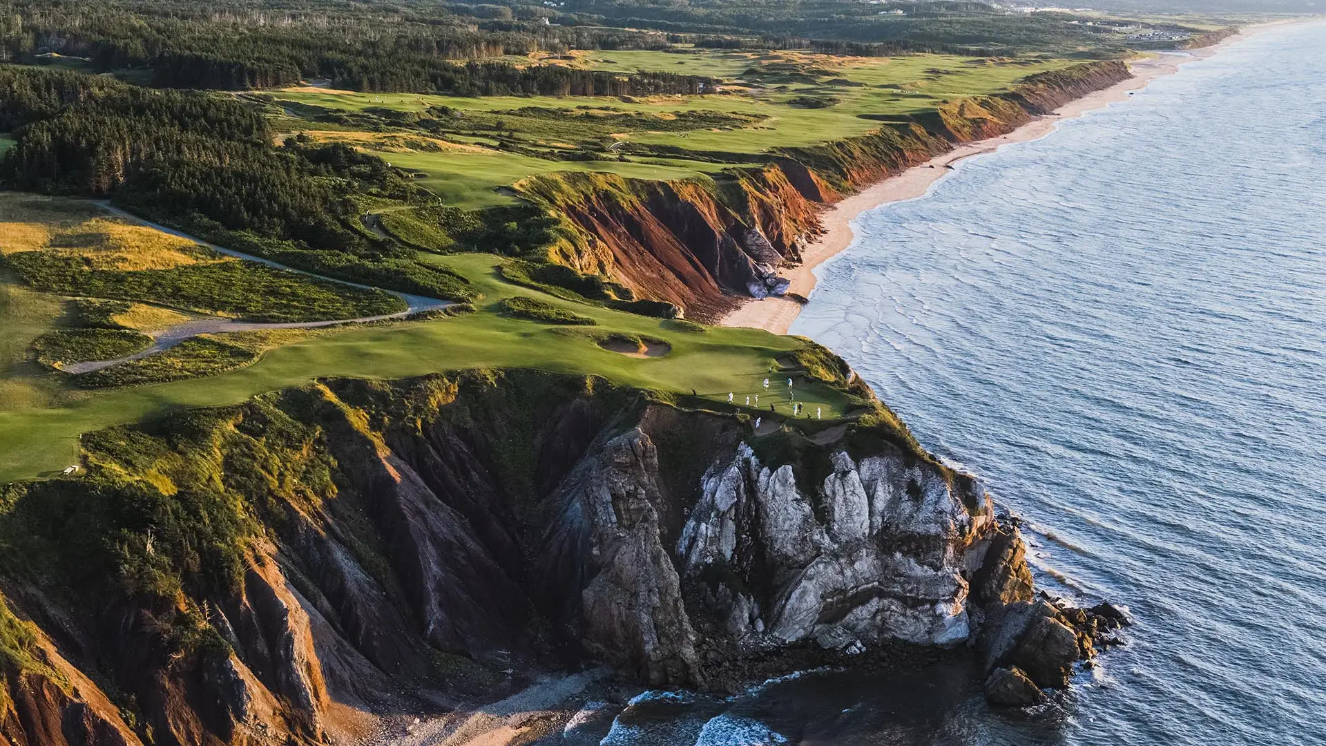 Dramatic aerial view of Cabot Cliffs golf course showing emerald fairways perched atop rugged red cliffs. The coastline stretches into the distance with pine forests inland and the Atlantic Ocean's blue waters meeting the rocky shore.