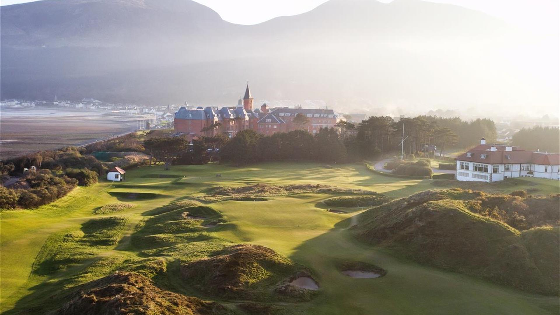 Morning view of Royal County Down Golf Course with dramatic sand dunes and undulating fairways in the foreground. A historic Irish town with prominent church spire sits nestled against misty mountains in the background. Golden morning light illuminates the links-style terrain while creating atmospheric shadows across the natural dunes.