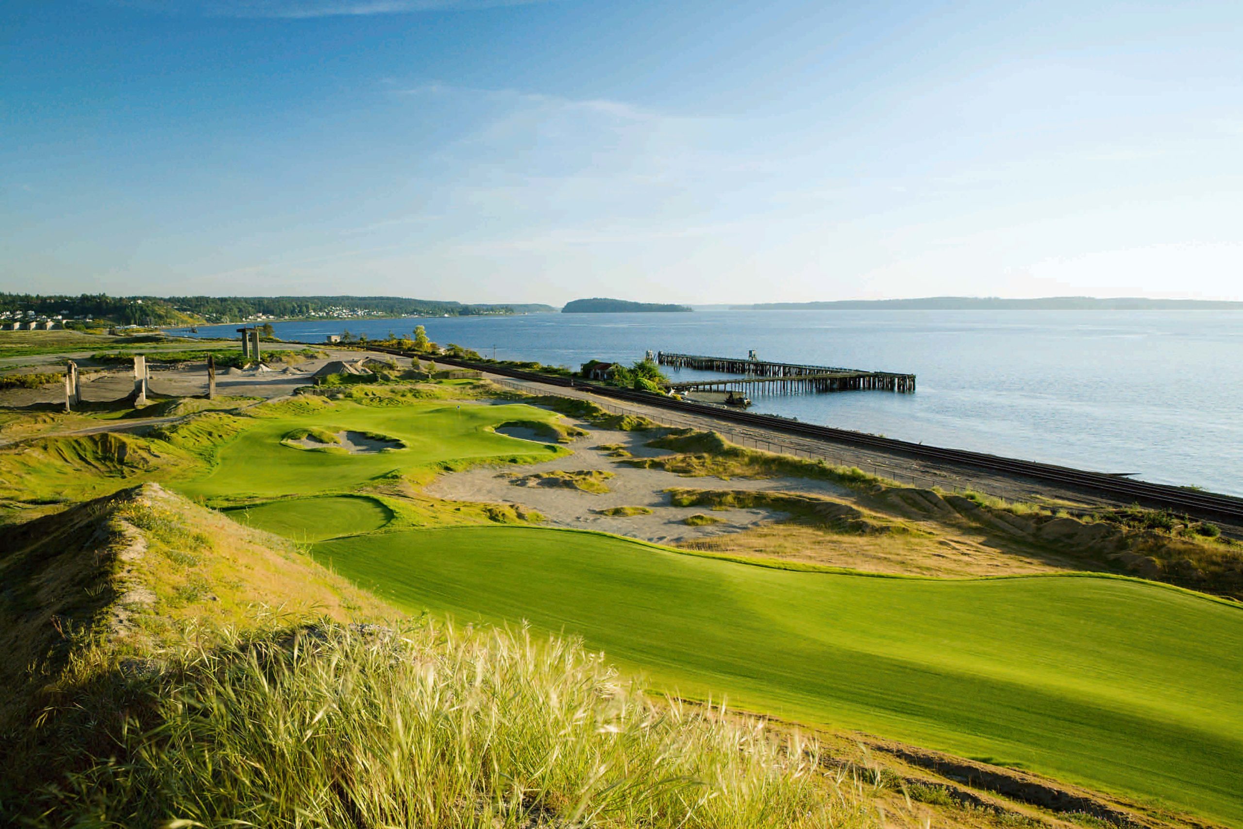 Aerial view of Chambers Bay Golf Course showing undulating fairways and sandy waste areas along Puget Sound. The course's natural contours and fescue rough create a striking contrast with the calm waters of the sound stretching to distant mountains.