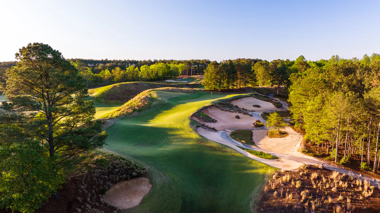 Aerial view of a golf hole at Tobacco Road featuring dramatic sand waste areas, undulating green fairways, and pine forest surroundings. The late afternoon sunlight creates striking shadows across the sculptured terrain and bunkers.