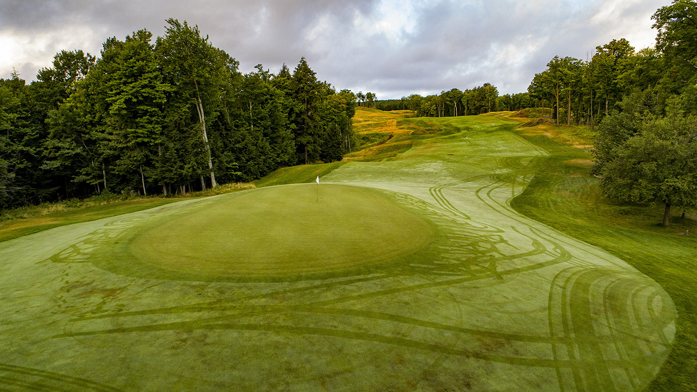 View of a large undulating green complex at Greywalls Golf Course, surrounded by dense forest. The green's contours are highlighted by early evening light, showing the dramatic movement in the putting surface.