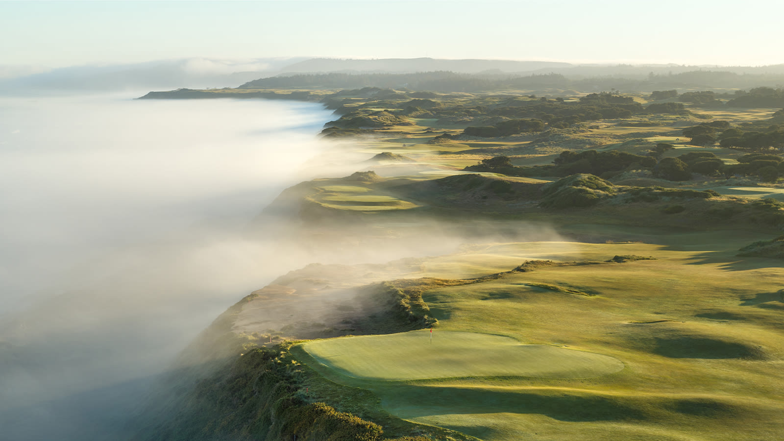 Aerial view of Bandon Dunes Golf Resort during a misty morning, showing emerald green fairways winding through natural dunes. Rolling fog dramatically spills over the coastal cliffs, creating a mystical atmosphere where the Pacific Ocean meets the rugged Oregon coastline.