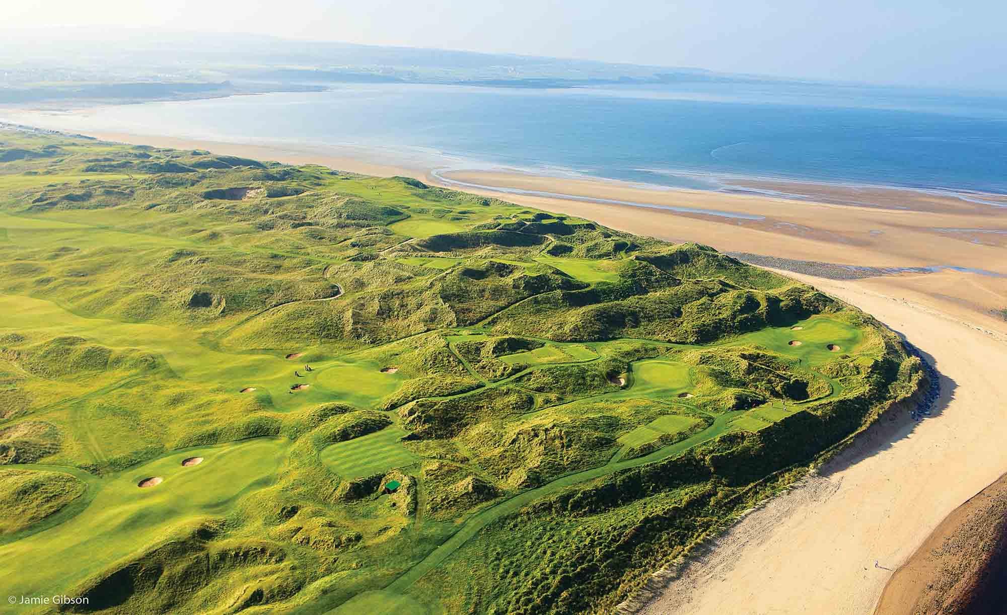 Evening view of Lahinch Golf Club's undulating links course with dramatic sand dunes in golden light. A historic Irish town with prominent church spire sits in the background against misty mountains, creating a quintessential Irish golf scene.Aerial view of Lahinch Golf Club showing emerald green fairways winding through natural sand dunes along a pristine beach. The dramatic Irish coastline stretches into the distance with turquoise waters meeting golden sand, while undulating links terrain creates a classic seaside golf landscape.