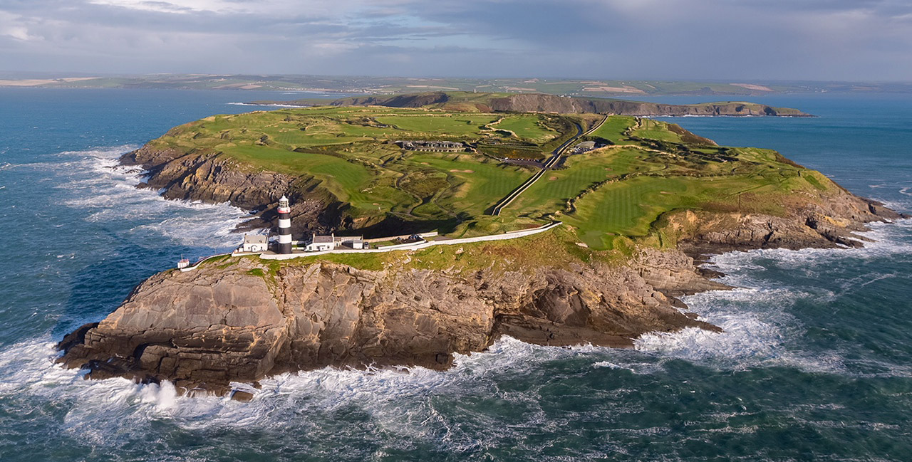 Aerial view of Old Head Golf Links in Ireland, showing a dramatic peninsula with emerald green fairways perched atop 300-foot cliffs. A distinctive black and white lighthouse stands at the point, surrounded by crashing Atlantic waves against rugged coastline.