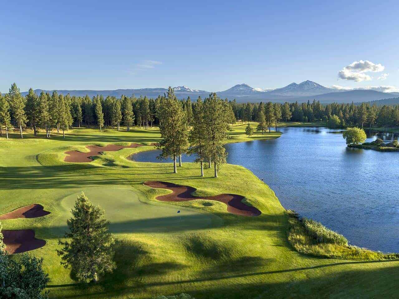 Aerial view of a lush green golf course with red sand bunkers, surrounded by pine trees and a lake, with snow-capped mountains in the background.