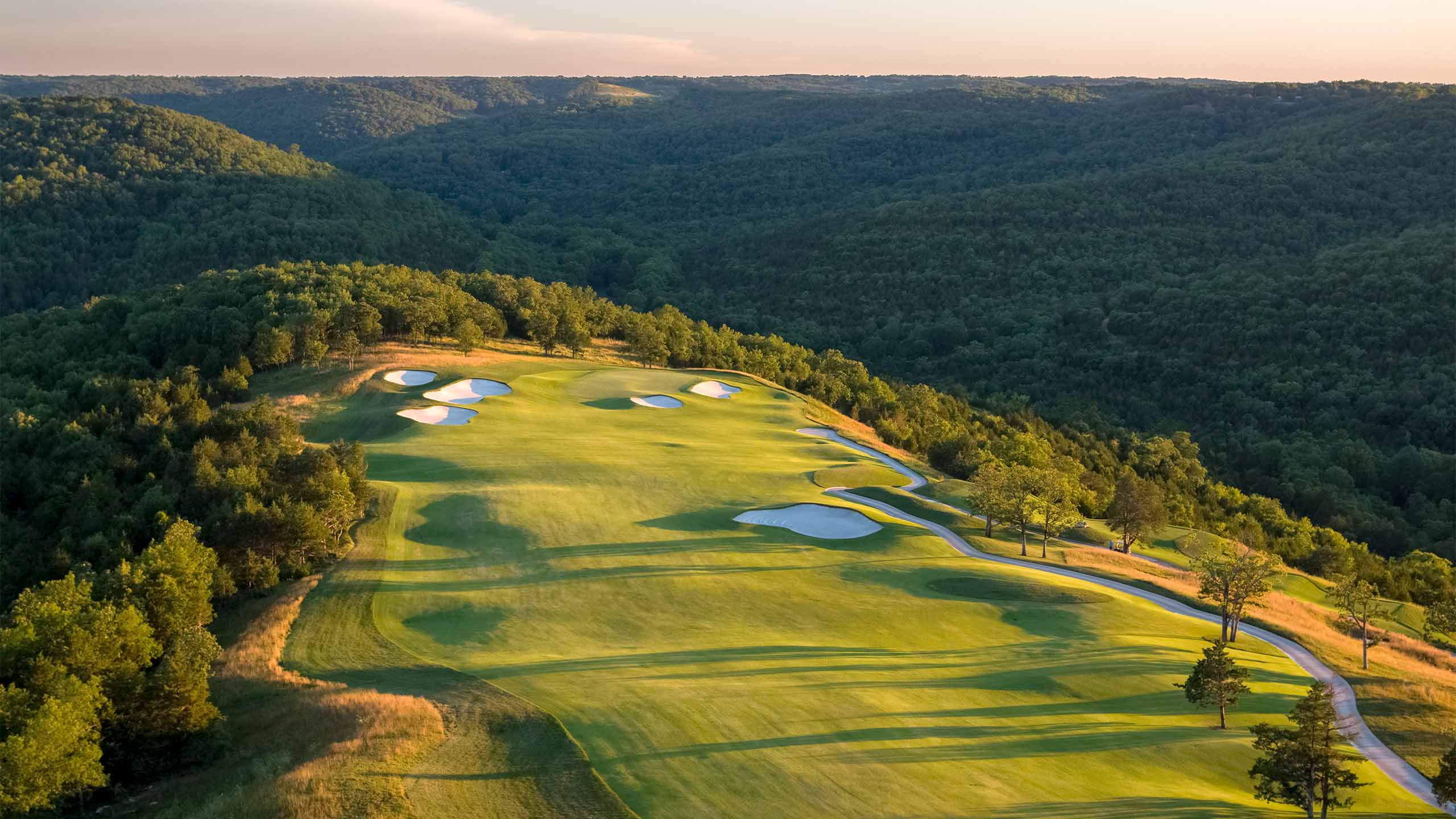 Aerial view of Payne's Valley Golf Course showing lush green fairways, white sand bunkers, and a winding cart path set against a backdrop of densely forested Ozark Mountains at sunset.