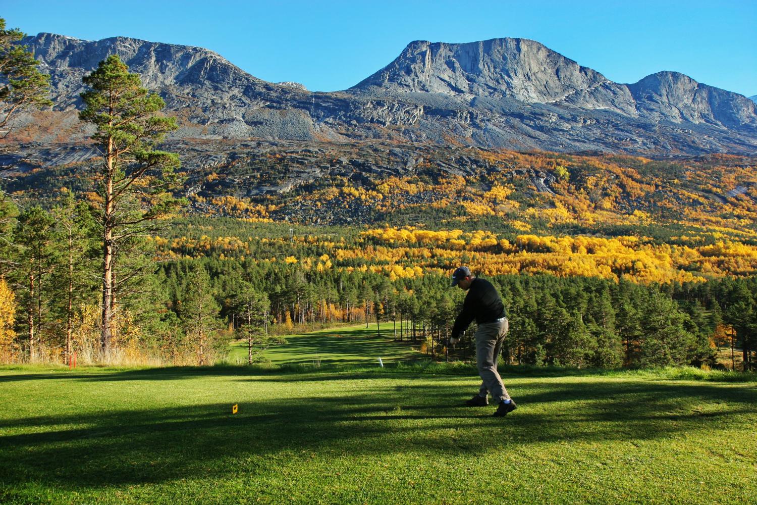 A golfer mid-swing on a tee box, with a backdrop of colorful autumn foliage and towering rocky mountains.